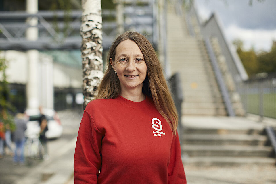 Stadtführerin Danica Graf von Suprise lächelt in die Kamera. Sie trägt einen roten Pulli mit dem Surprise Logo und steht auf der Dreirosenmatte
